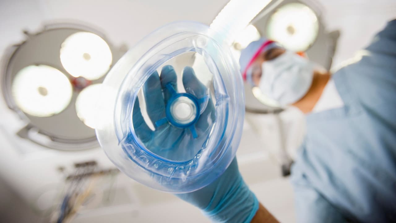 A provider lowers an anesthesia mask — patient's-eye view under operating room lights.